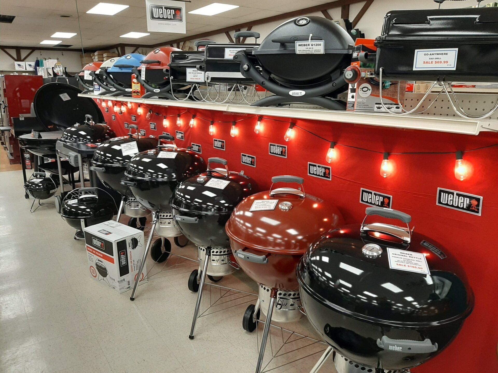 Row of Weber charcoal grills on display in a store, various colors, red background.