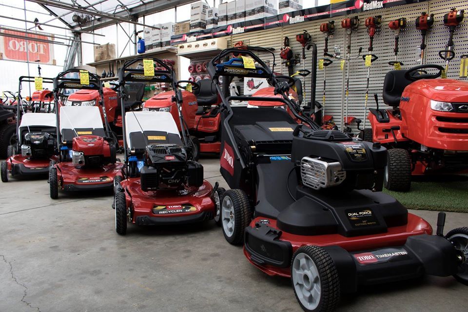 Row of red and black lawnmowers displayed in a hardware store, with various models and styles visible.