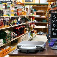 Inside a store: shelves stocked with items, a scale, and a chalkboard sign on the counter.