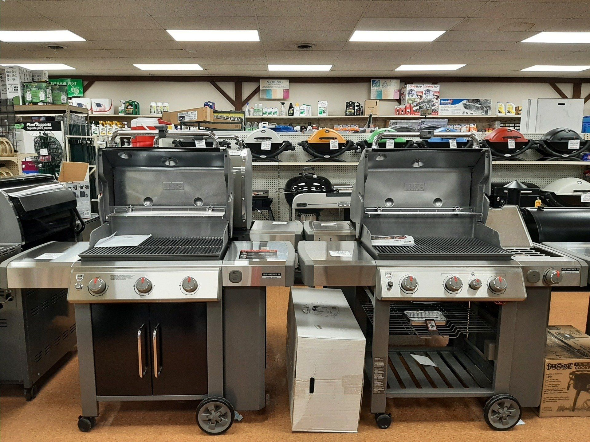 Two gas grills displayed in a hardware store, featuring open lids, stainless steel surfaces, and gray cabinets.