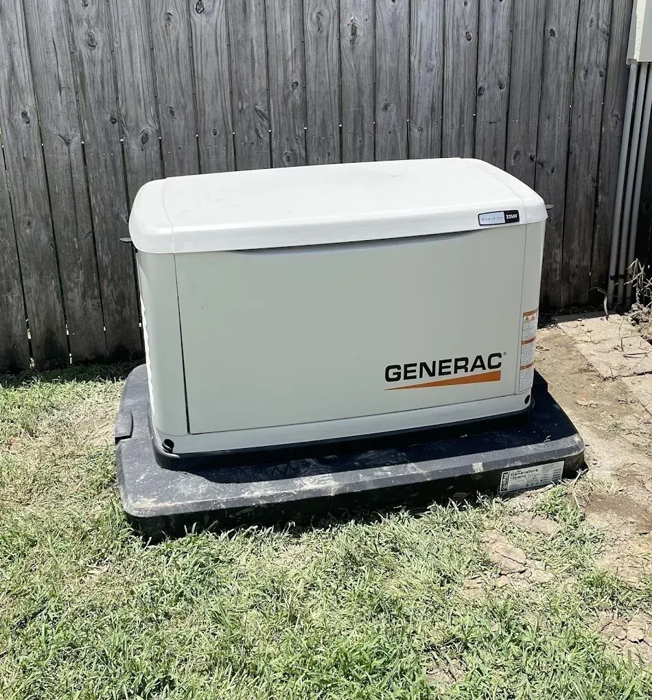 A generator is sitting in the grass in front of a wooden fence.