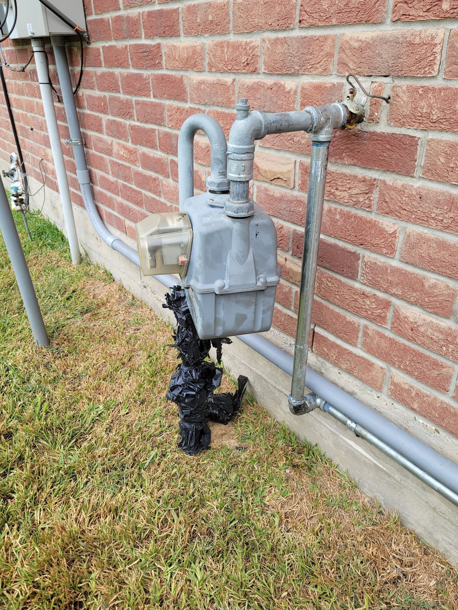 A gas meter is sitting on the grass next to a brick wall.