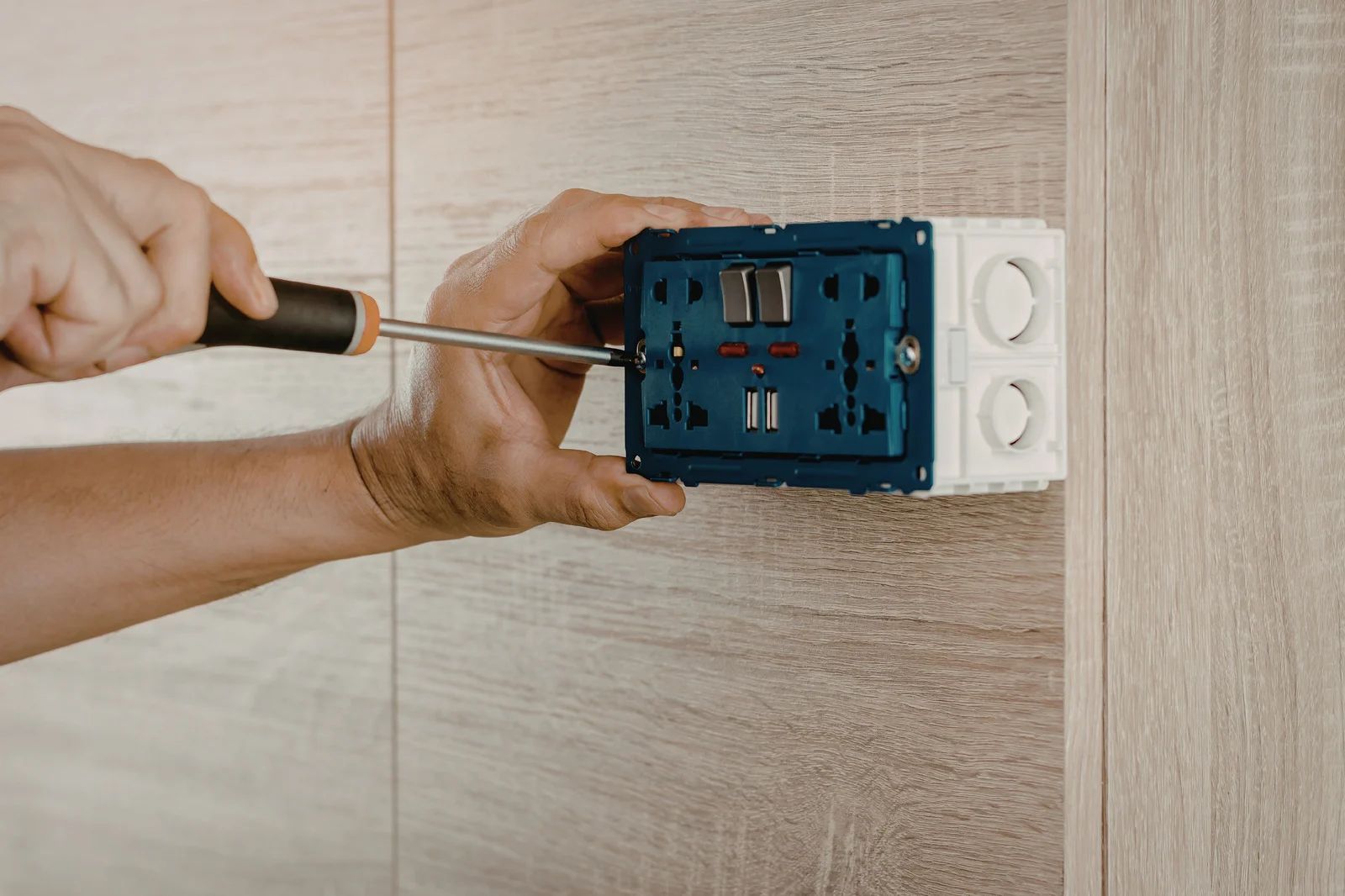A person uses a screwdriver to mount a blue electrical wall outlet into a wooden panel.