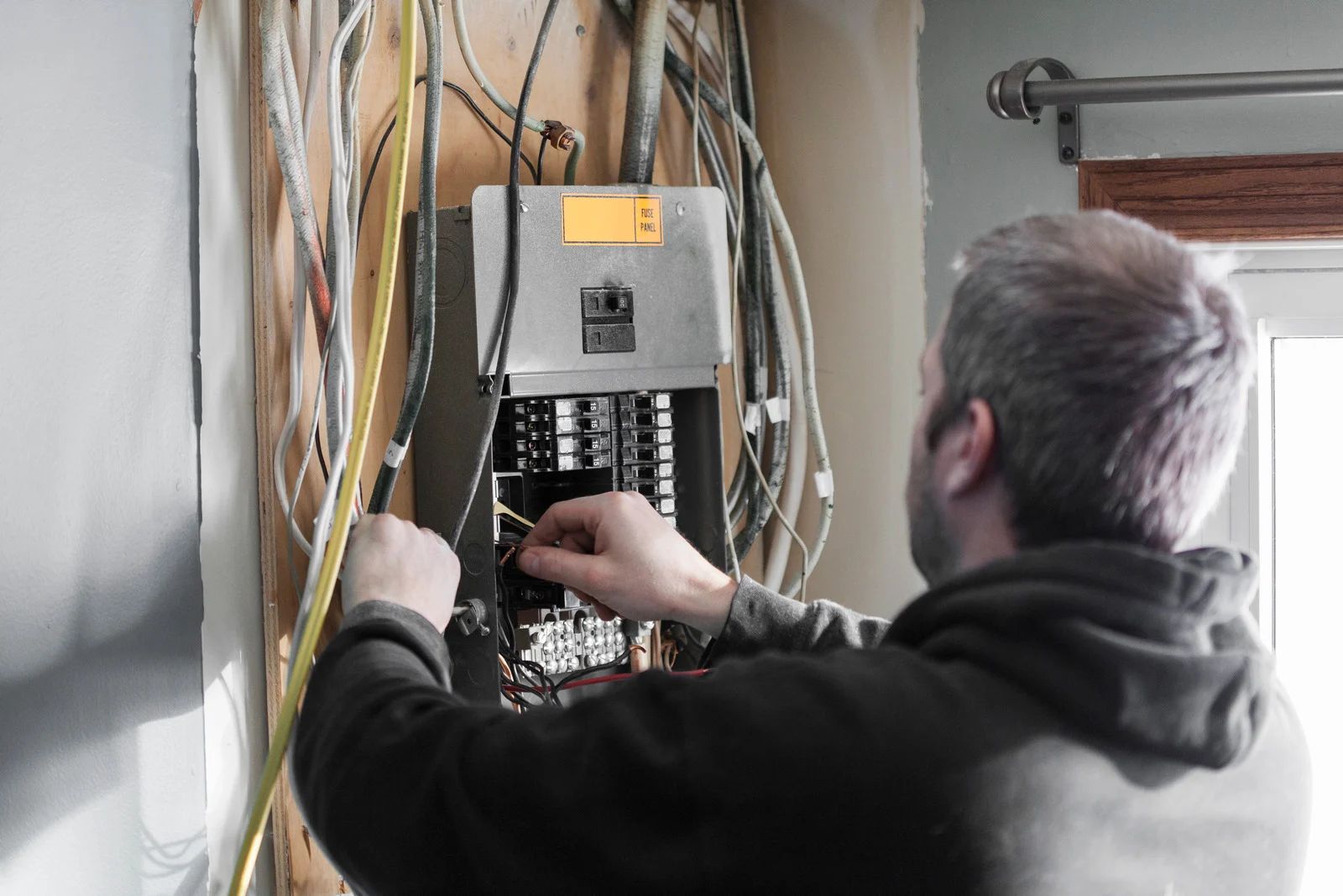 Man working on an electrical panel, surrounded by wires, inside a room.