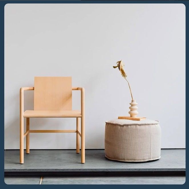 Una sedia in legno chiaro accanto a un piccolo pouf con fiori secchi, contro una parete bianca.