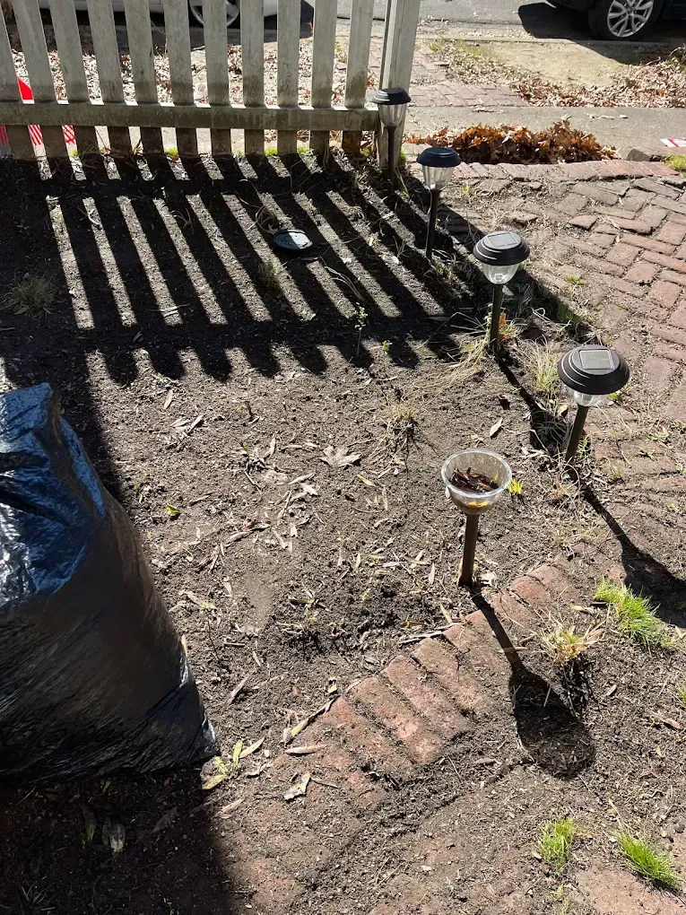 Garden path with solar stake lights beside a white fence, showing shadow patterns on dirt and brick paving.