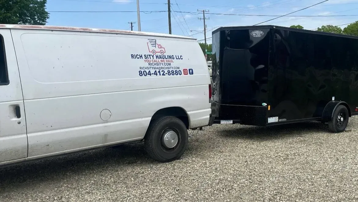 White work van towing a black enclosed trailer parked on gravel under a blue sky