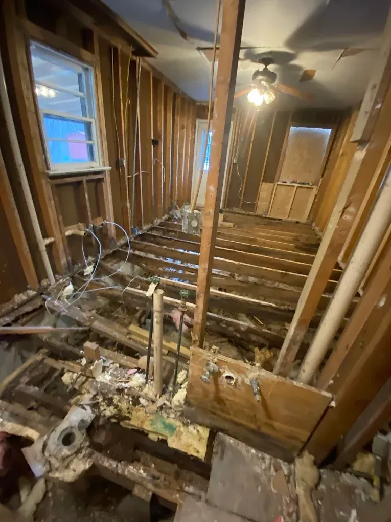 Interior of a stripped house under renovation, with exposed wall studs, floor joists, and debris.