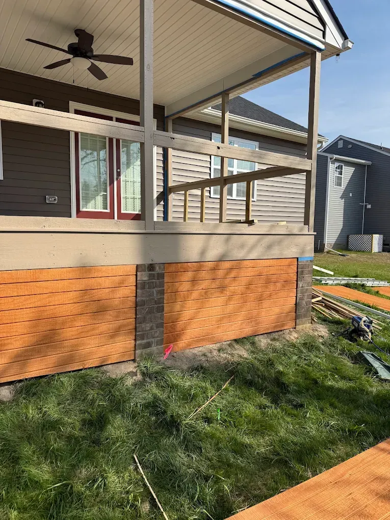 Covered porch with beige siding, brick skirt, and a grassy yard in front of a house.