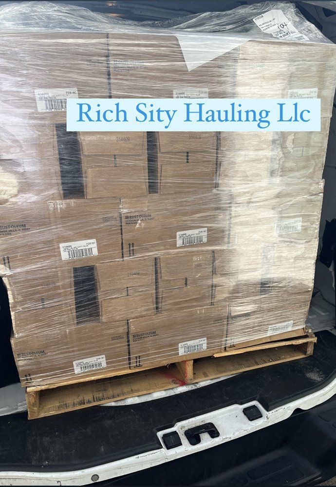 Shrink-wrapped cardboard boxes on a wooden pallet in a truck bed, labeled “Rich City Hauling LLC.”