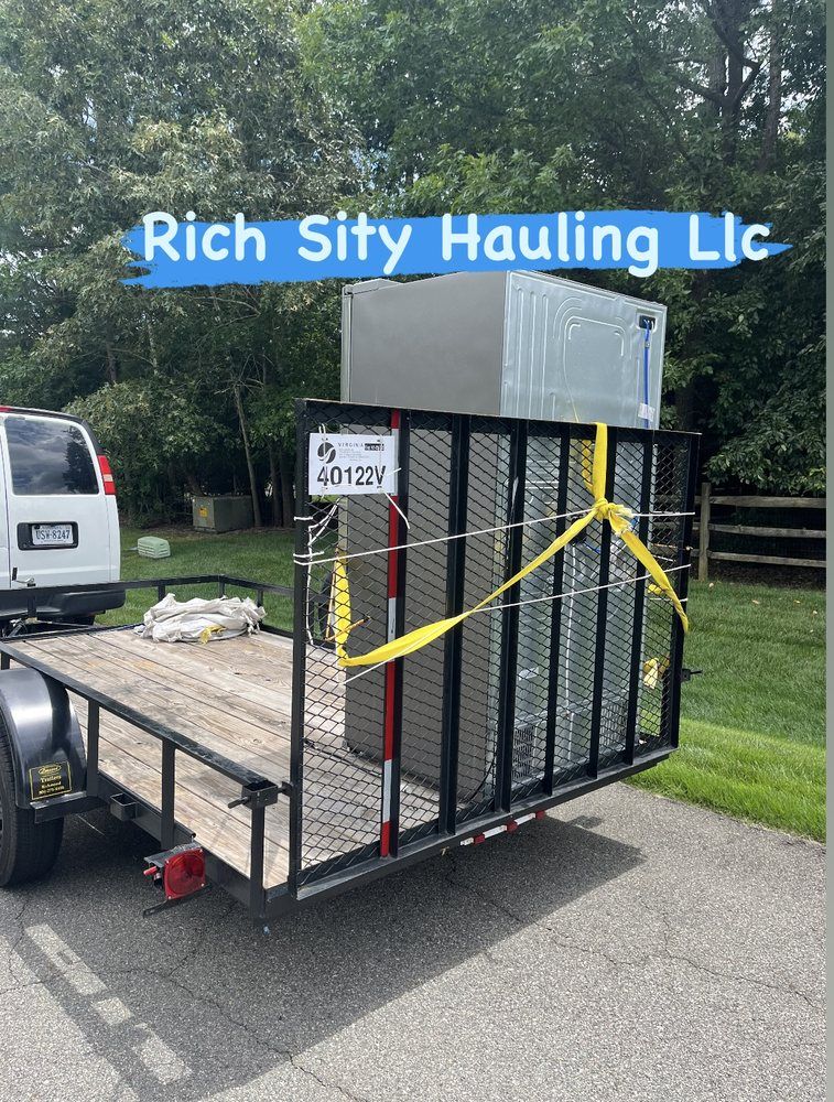 Utility trailer with metal cage, yellow straps, and appliance load parked by a roadside fence.