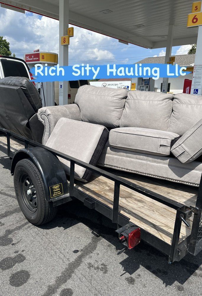 Black pickup truck hauling a beige sectional sofa on a flatbed trailer at a parking lot.