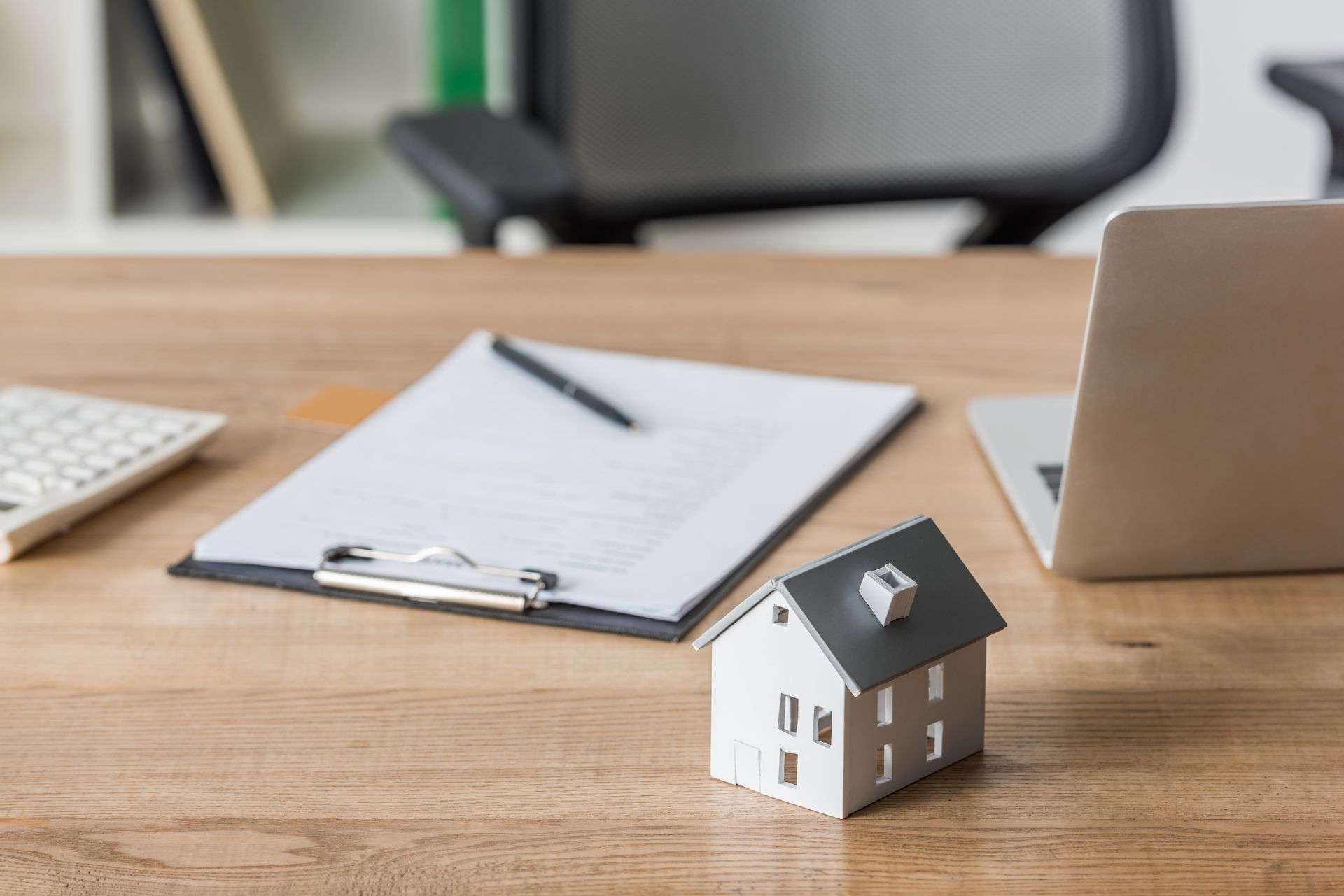 Wooden desk with a toy house, clipboard with pen, laptop, and calculator; office setting.