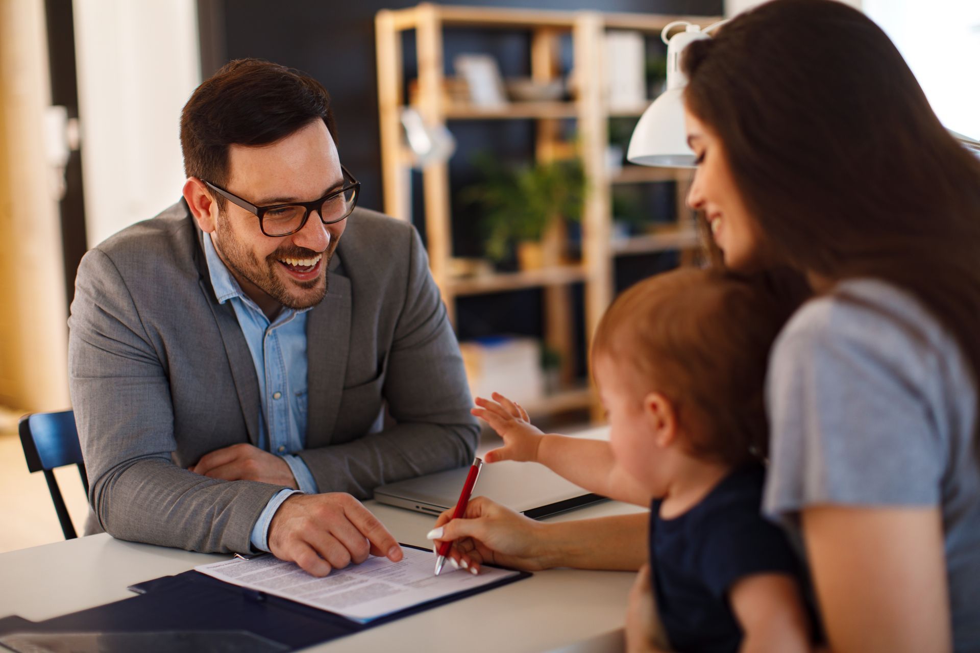 Man in suit smiles at woman signing document with child in lap.