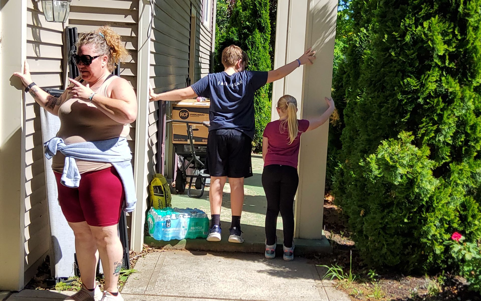 Three people pose outdoors near a building with outstretched arms. The person on the left wears sunglasses.