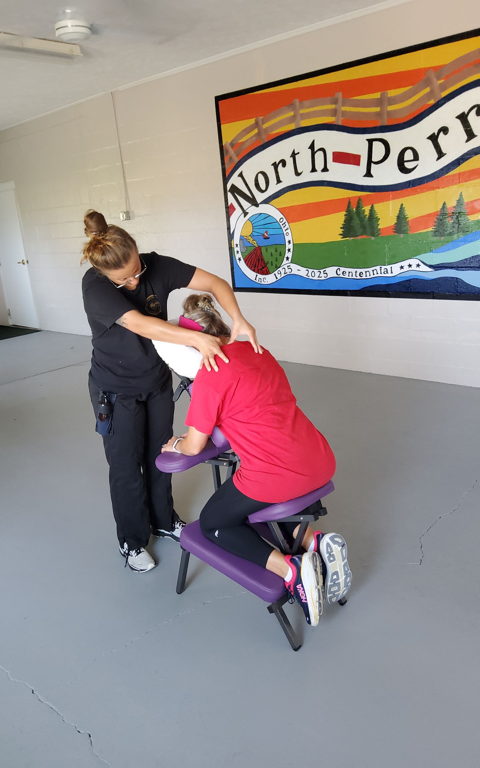 Woman giving a seated shoulder massage to another woman, in front of a painted wall mural.