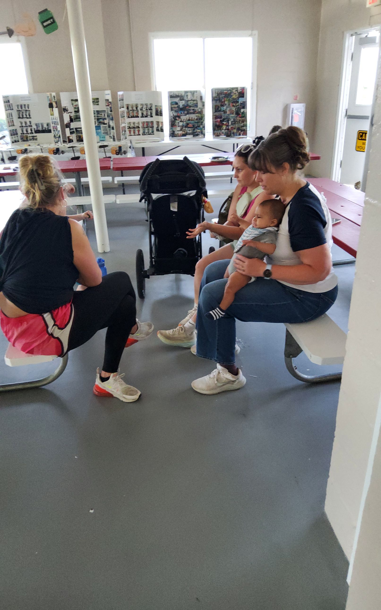 Four people, including a person holding a baby, seated in a room, conversing near a stroller and a table.