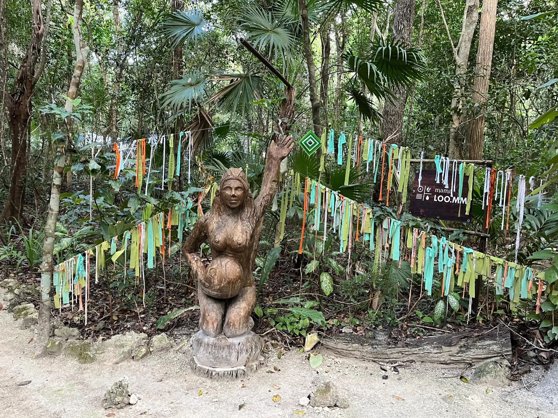 Estatua de piedra de una mujer en un entorno forestal, con los brazos en alto, rodeada de cintas de colores.