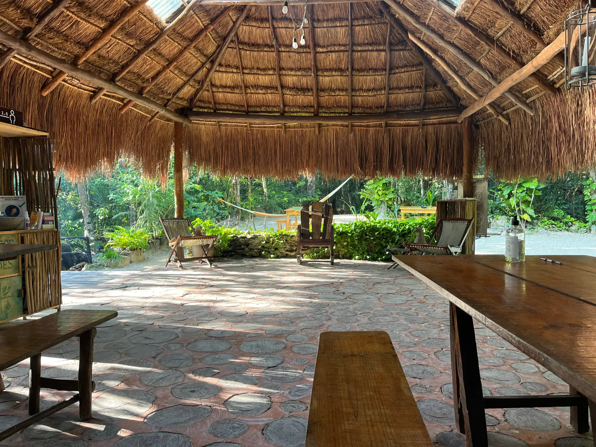 Open-air thatched-roof structure with wood furniture and view of greenery.