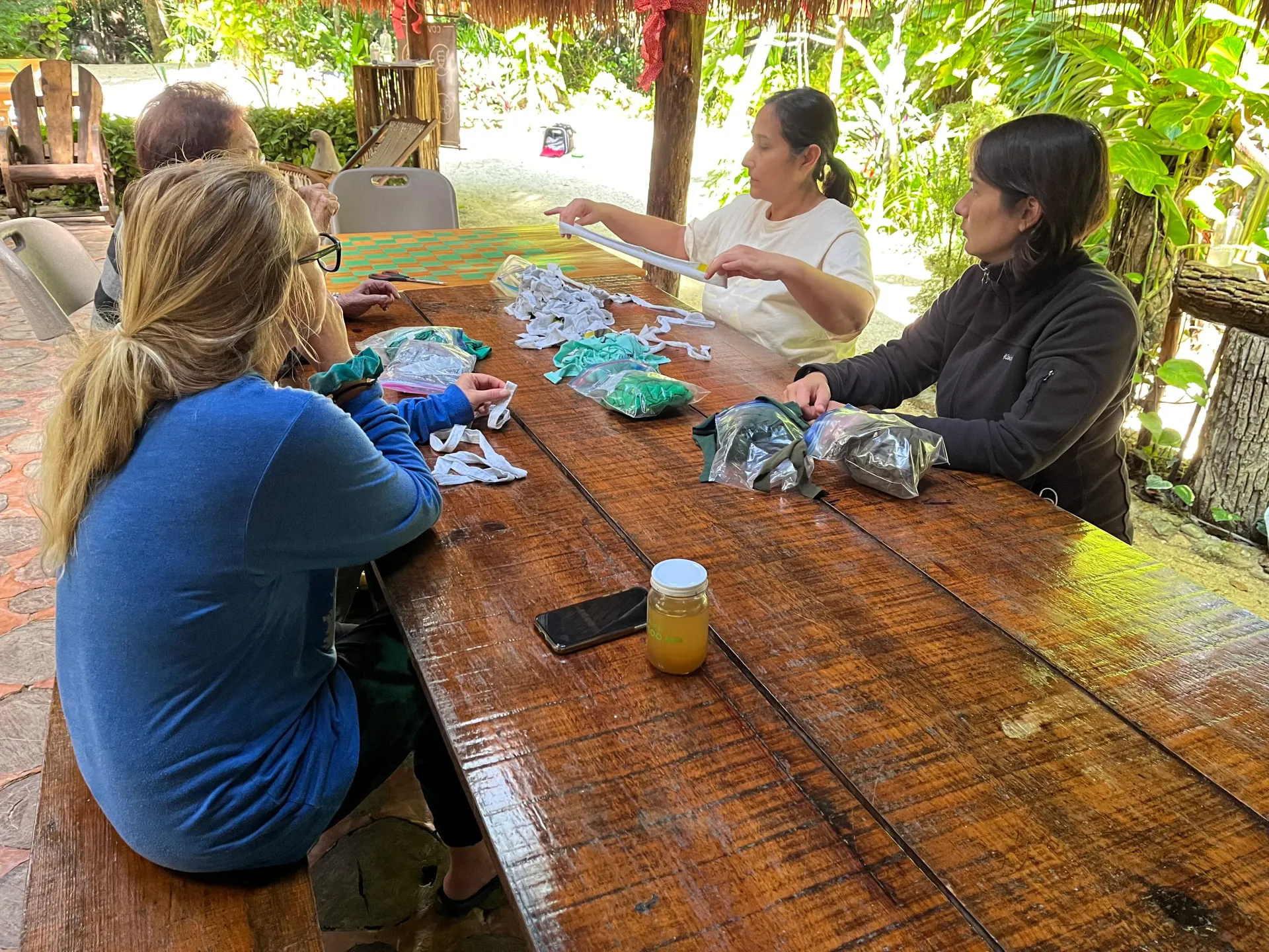 La gente se reúne alrededor de una mesa de madera y trabaja en artesanías al aire libre, bajo un techo de paja.