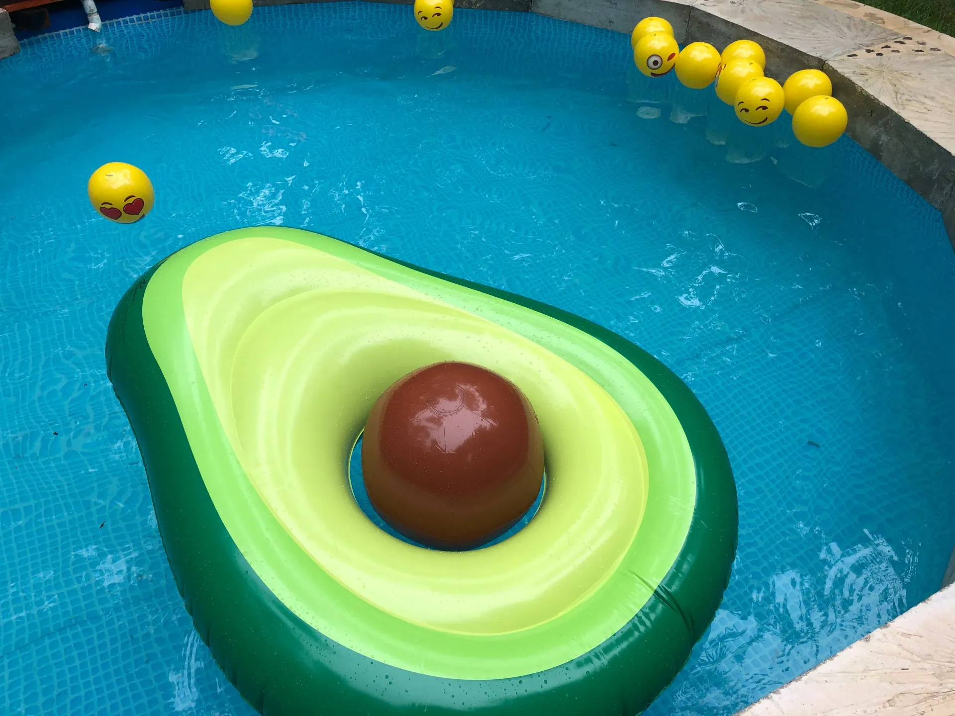Avocado pool float in a blue pool with yellow smiley face balls.