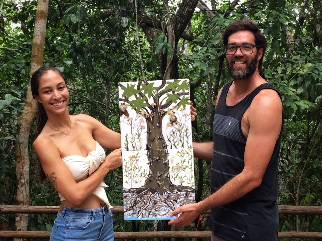Una mujer y un hombre sostienen una obra de arte de un árbol en un bosque. Ambos sonríen y visten ropa informal.