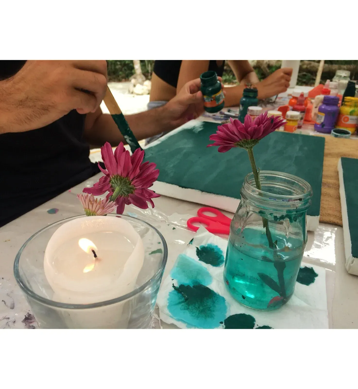 Persona pintando lienzo con pintura verde, flor, vela y jarra de agua sobre mesa.