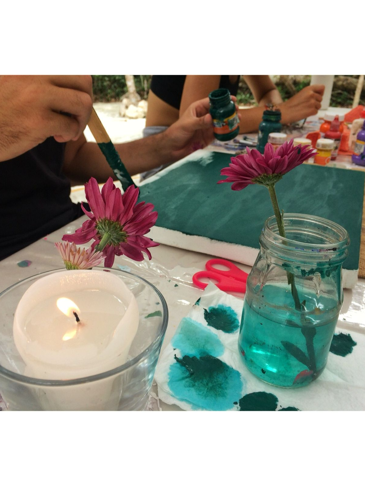 Personas pintando con flores y velas sobre una mesa. Una persona pinta verde sobre un lienzo; una flor rosa en un frasco.