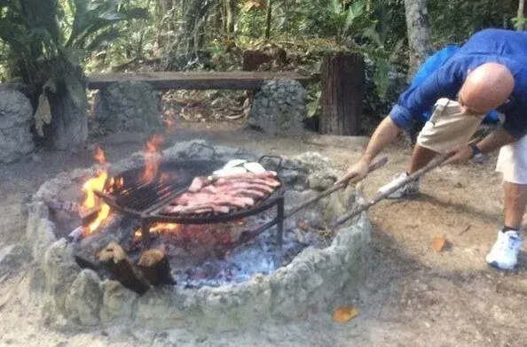 Hombre asando carne sobre una fogata en un pozo de piedra, al aire libre.