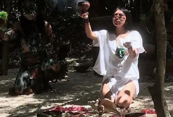 Mujer vestida de blanco, arrodillada, sosteniendo un cuenco durante un ritual en un entorno forestal.
