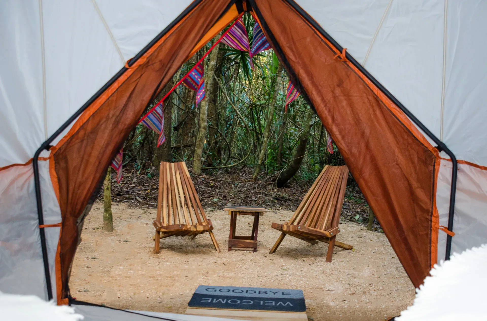 Dentro de una tienda de campaña, dos sillas y una pequeña mesa miran hacia la vegetación con un tapete de bienvenida