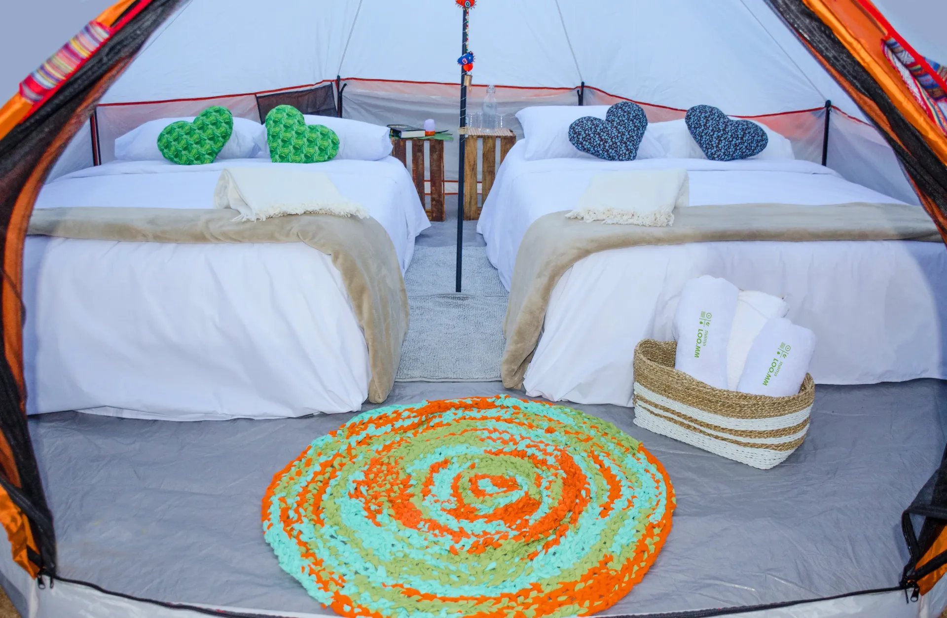 Dentro de una tienda de campaña: dos camas con almohadas en forma de corazón, una alfombra y una canasta.