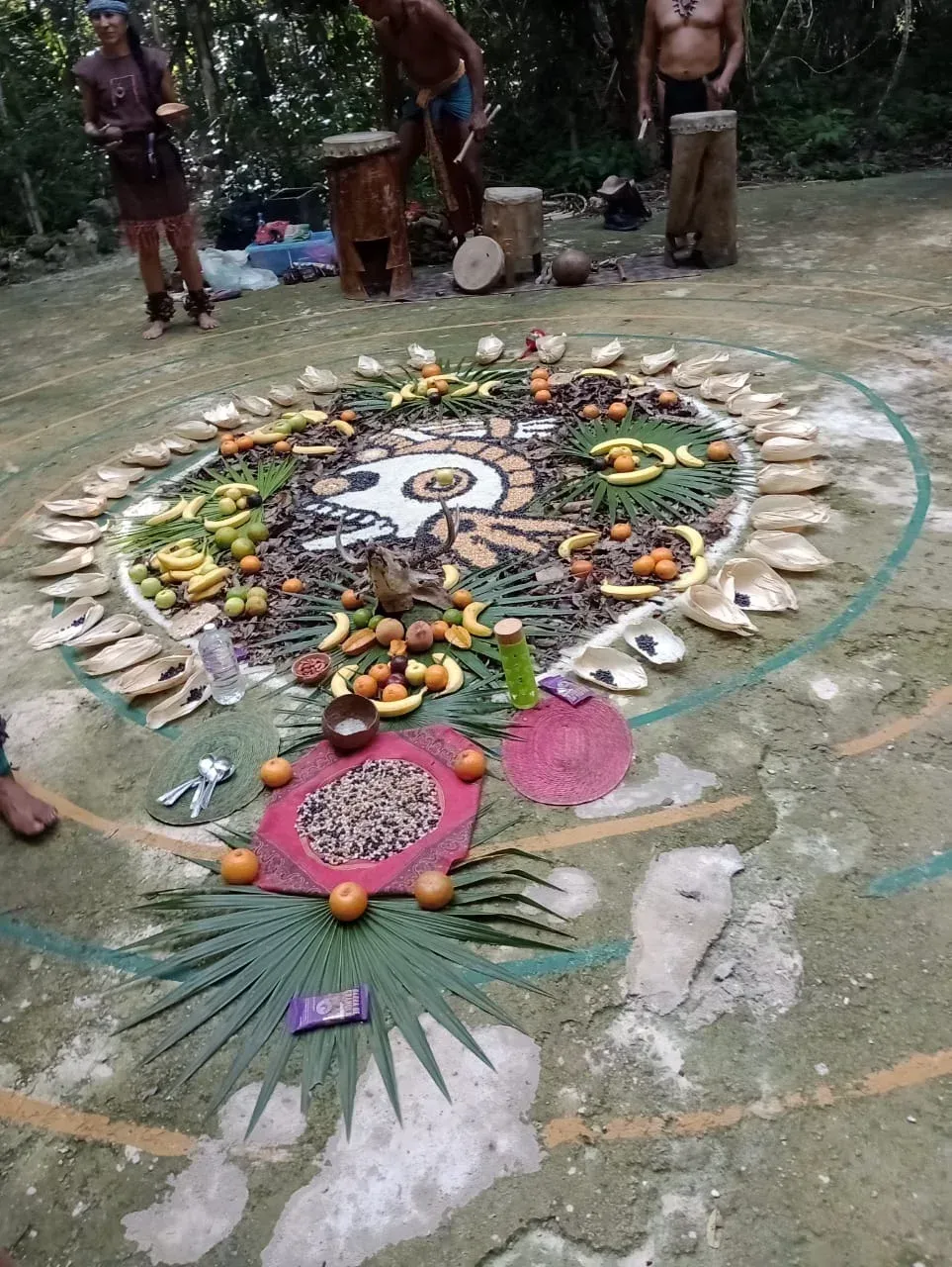 Reunión ceremonial con gente, mandala de comida y músicos en un entorno forestal.