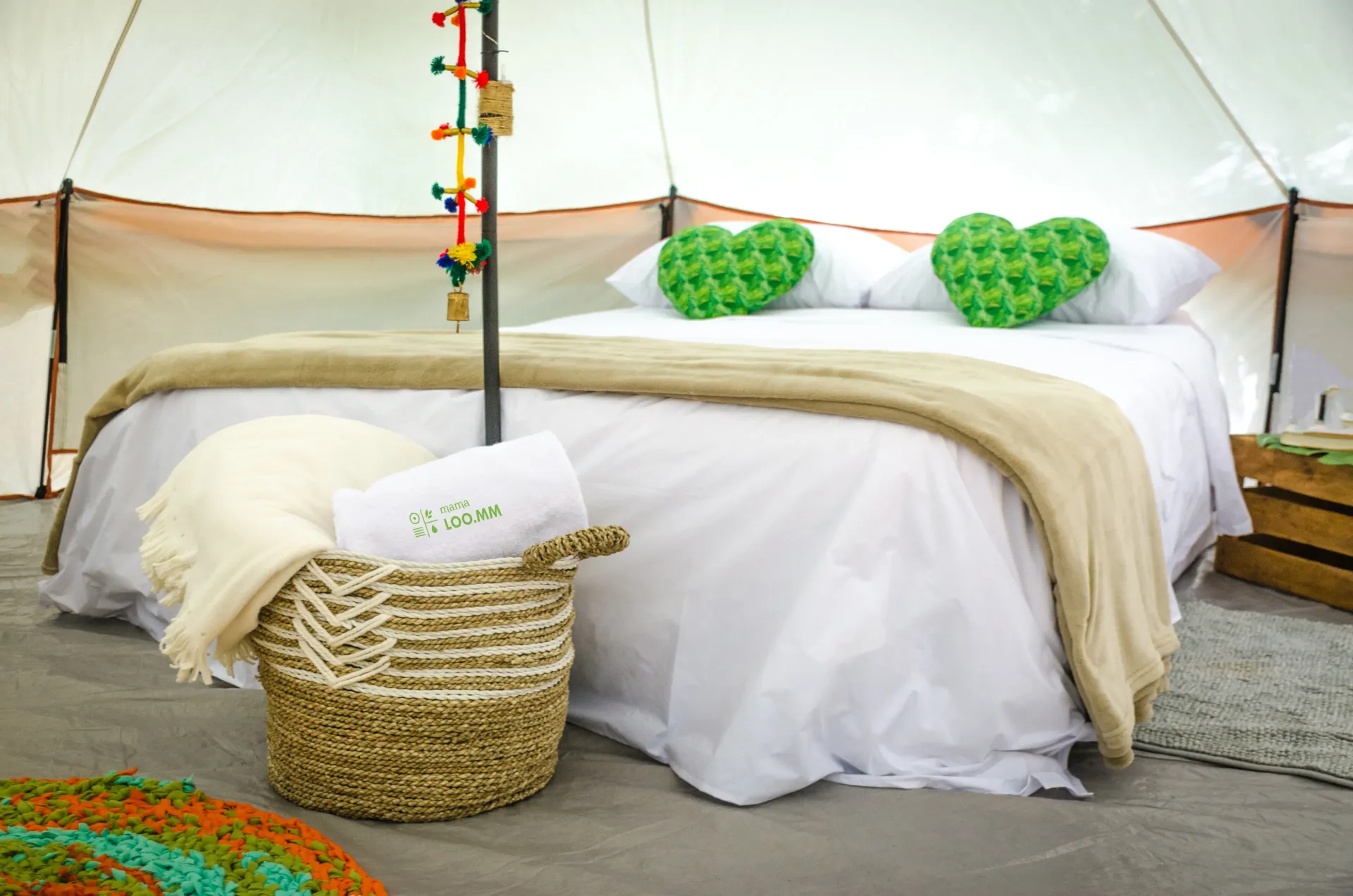 Dentro de una tienda glamping, una cama con almohadas en forma de corazón, una canasta con toallas y decoraciones coloridas.