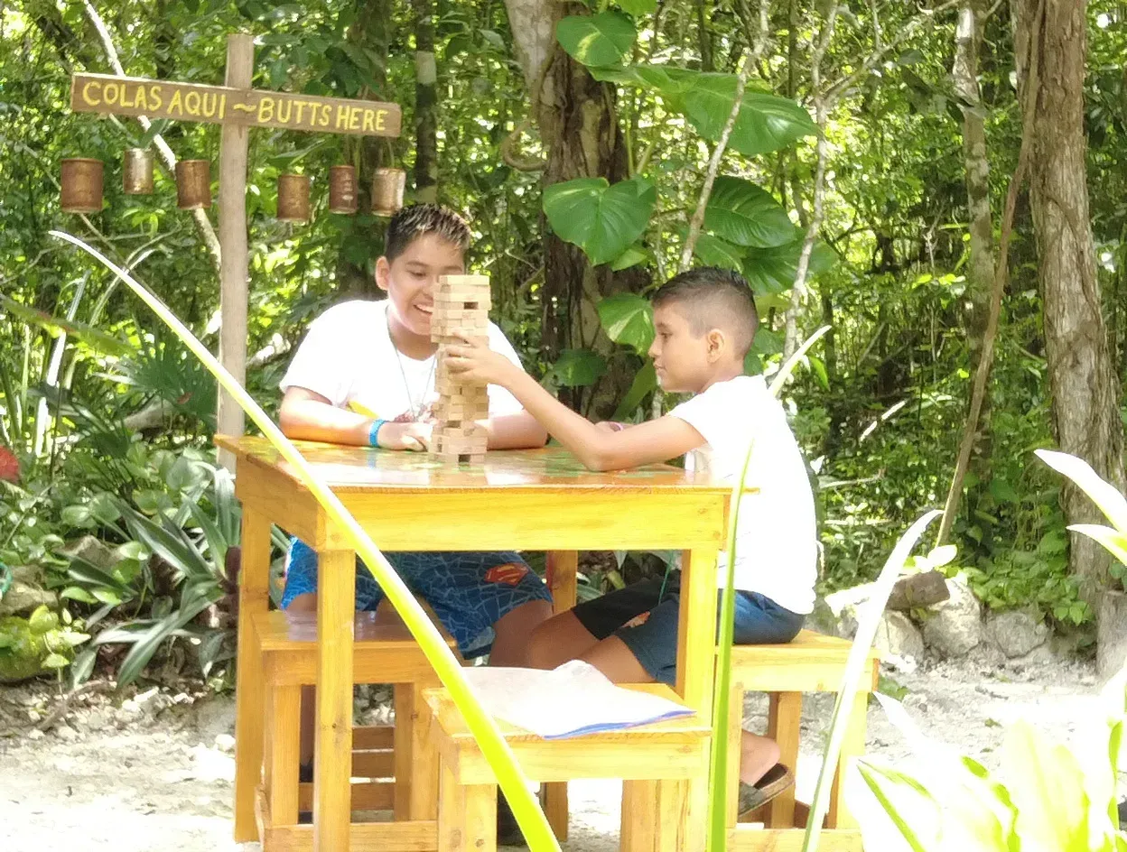 Dos niños jugando Jenga en una mesa de madera al aire libre con árboles al fondo.