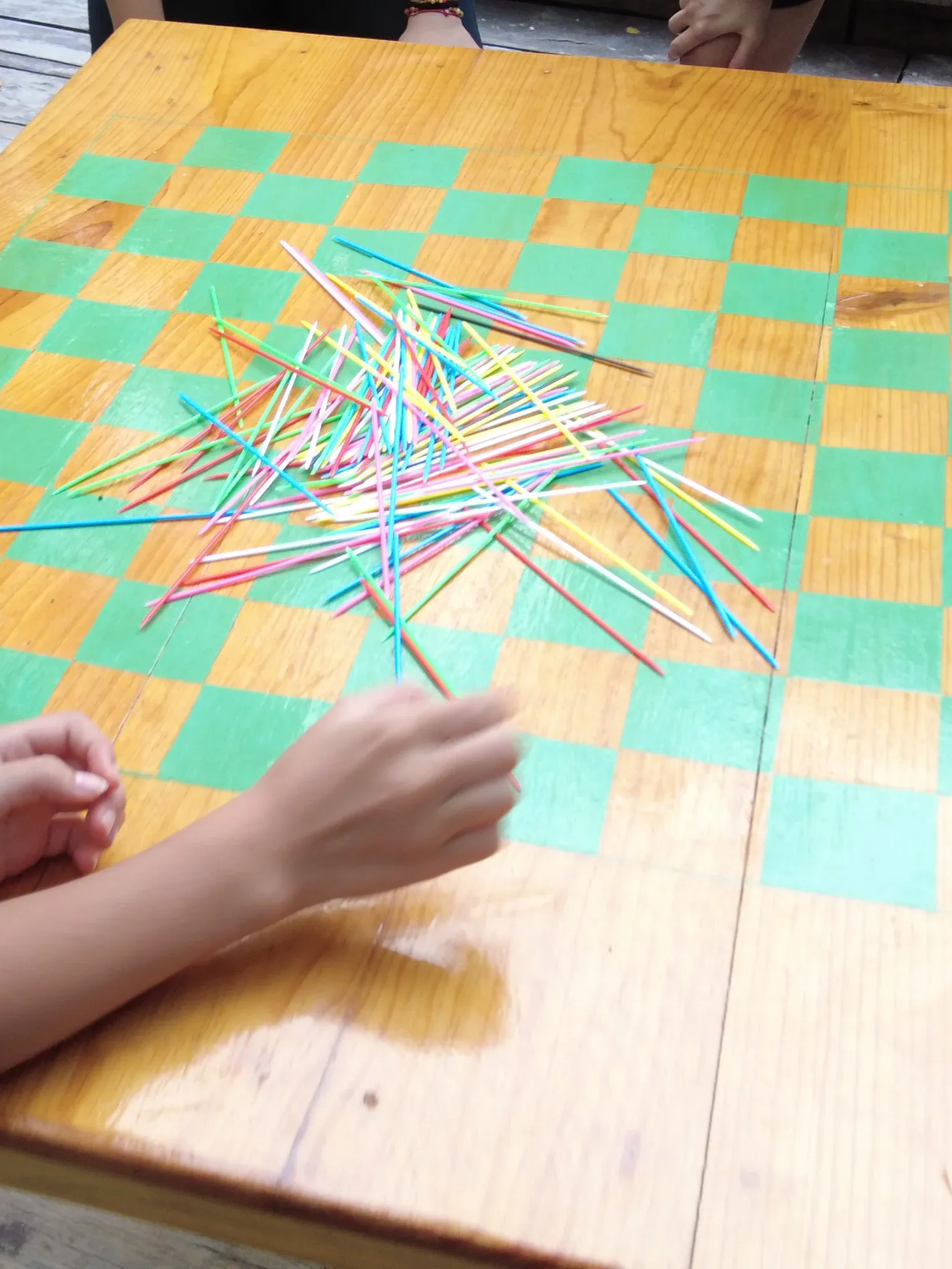 Persona jugando a los palitos en una mesa a cuadros. Palitos de colores dispersos, con una mano sujetando uno.
