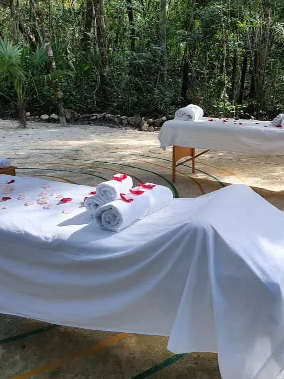 Mesas de masaje al aire libre con mantelería blanca, toallas enrolladas y pétalos de rosa en una zona boscosa.