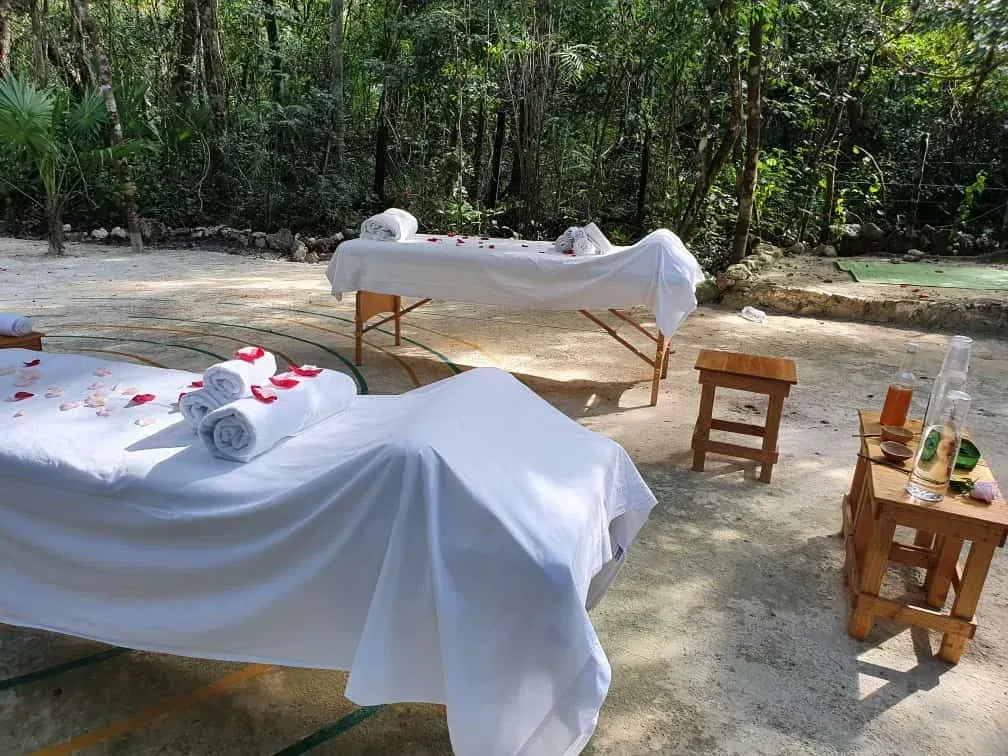 Two massage tables with white linens and rose petals outdoors near trees.