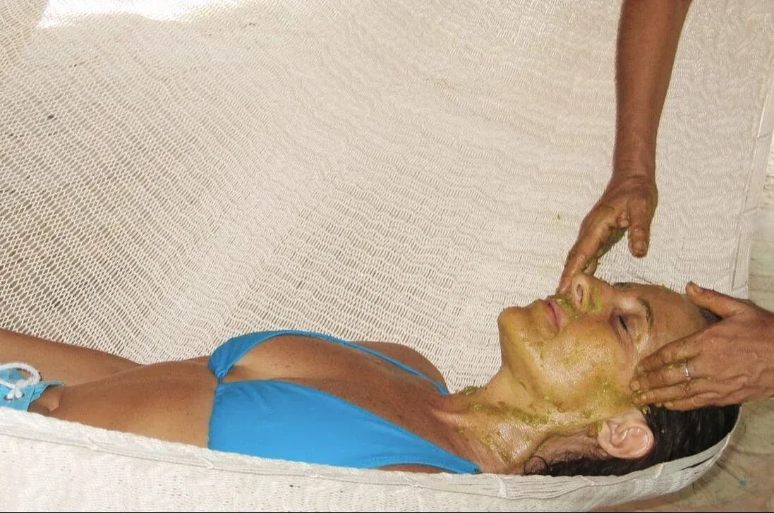 Mujer recibiendo tratamiento facial, acostada sobre una superficie blanca, con las manos aplicando una mascarilla.