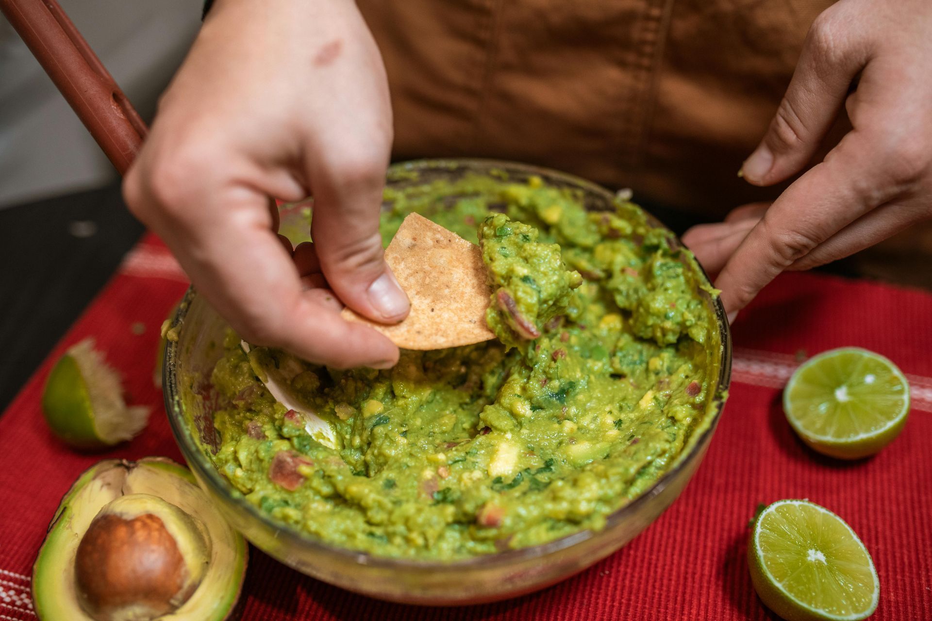 Persona sumergiendo un totopo en un tazón de guacamole. Limones y aguacate sobre una superficie roja.