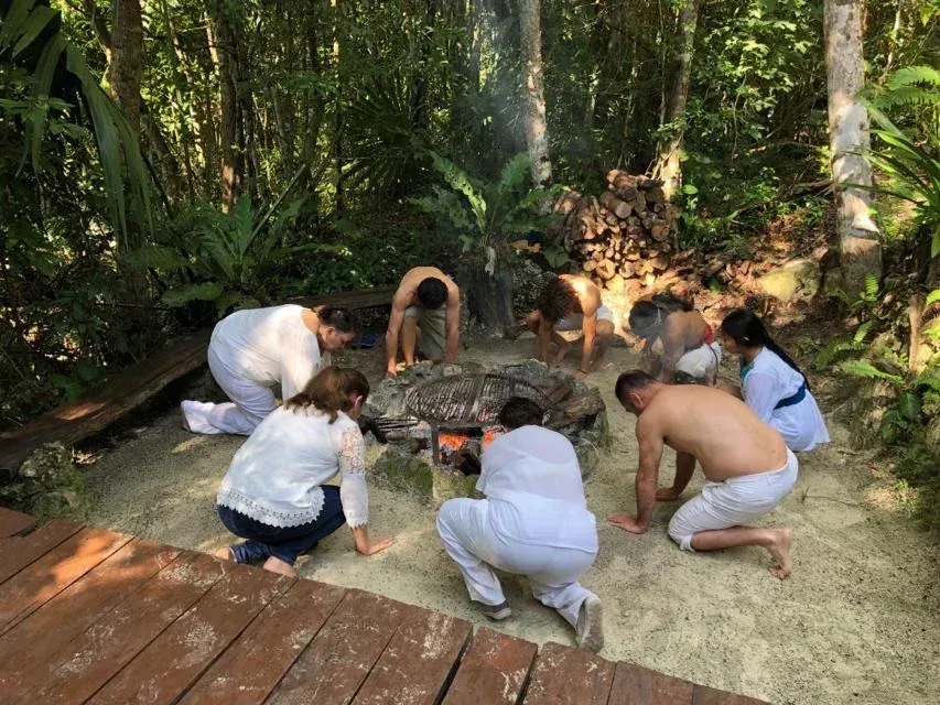 Un grupo de personas vestidas de blanco, arrodilladas alrededor de una fogata en un frondoso bosque.