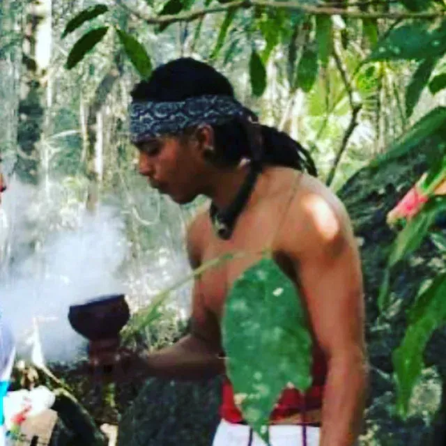 Hombre en la naturaleza, sosteniendo un recipiente humeante. Lleva un pañuelo y un collar, y está parcialmente sin camisa.