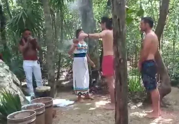 Personas en una jungla participando en una ceremonia. Una mujer está siendo rociada con agua, mientras otros observan.