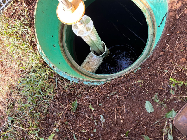 Open green manhole with pipes and water inside, surrounded by dirt and grass