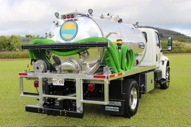 White vacuum truck with green hoses parked on grass in a field, with hills in the background