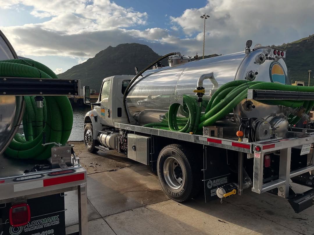 Silver vacuum truck with green hoses parked outdoors near a mountain under a cloudy sky