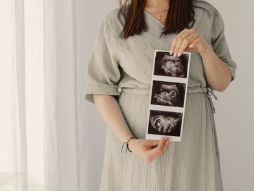 Pregnant person in a gray dress holding ultrasound printouts in front of their torso