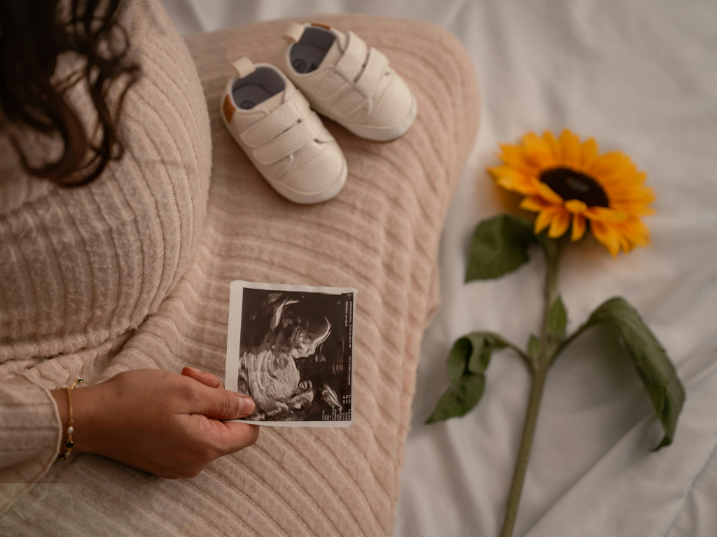 Ultrasound photo held on a beige blanket beside baby shoes and a sunflower vase