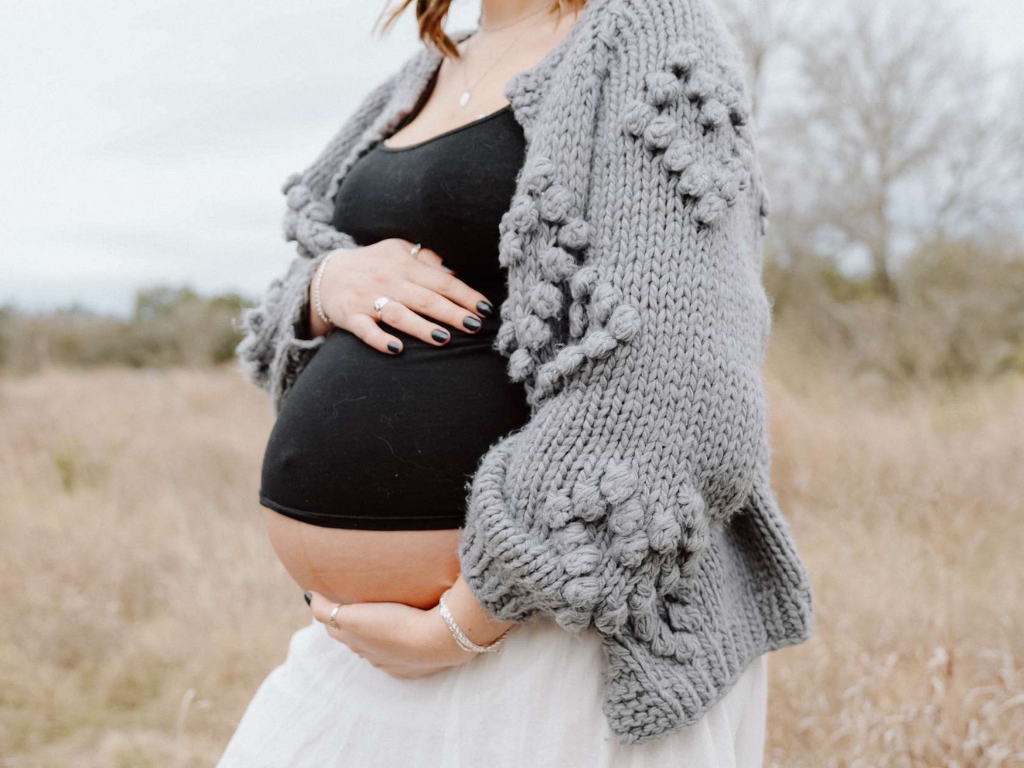 Pregnant person outdoors, holding a belly with hands in a gray sweater and black top.