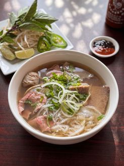 Bowl of pho with herbs and lime wedges on a table, with a small chili sauce dish nearby